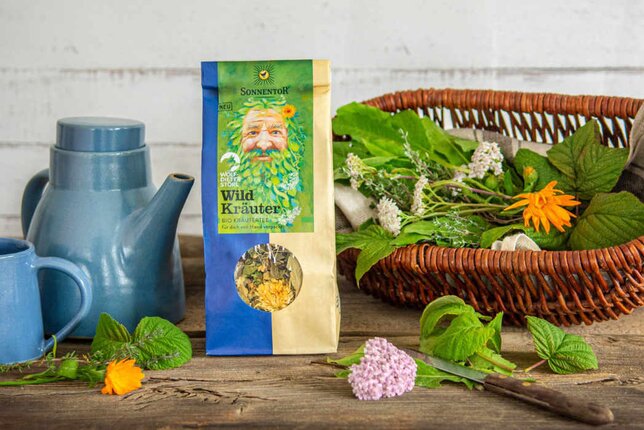 Photo of a package of wild herbal tea. Next to it is a basket with fresh herbs. | © SONNENTOR