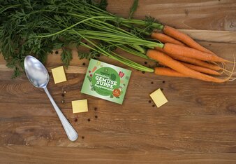 DAs Bild zeigt ein Päckchen Zuckerfreie Gemüsesuppe aus der Vogelperspektive aufgenommen, daneben liegen 3 golden verpackte Suppenwürfen, ein Löffel und ein paar Karotten mit Grün dran. | © SONNENTOR