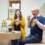 Das Foto zeigt Hannes und die Mitarbeiterin Theresa beim Trinken des Löwenzahnwurzelkaffees, beide sehen sehr fröhlich aus. Vor ihnen am Tisch steht die Kanne mit dem Kaffee und einige Dosen des Pulvers daneben aufgestapelt. | © SONNENTOR