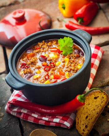 Auf dem Foto ist ein schwarzer Topf mit Chili con Carne auf einem rot kariertem Küchentuch zu sehen. Daneben sind Paprika und Brot zu sehen. | © SONNENTOR