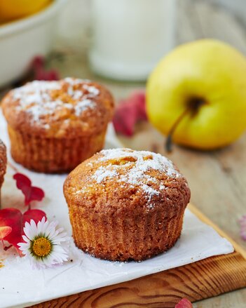 Auf dem Foto sieht man zwei Muffins. Daneben liegt ein Gänseblümchen und dahinter ist ein Apfel zu sehen. | © SONNENTOR