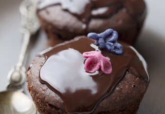 Foto von zwei Bananen-Kakao-Muffins auf einem Teller. Die Muffins sind mit Schokolade übergossen und mit Blüten garniert. | © SONNENTOR