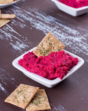 Auf dem Foto ist ein Schälchen mit Rote Rüben Dip zu sehen. Darin steckt ein Cracker. Auch vor der Schüssel liegen zwei Cracker. | © SONNENTOR