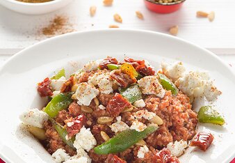 Auf dem Foto ist ein Teller mit Couscous-Tomaten-Pfanne zu sehen. Darüber ist Schafkäse verteilt. Im Hintergrund sieht man Pinienkerne. | © SONNENTOR