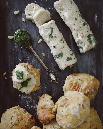 Auf dem Foto sieht man mehrere Tomatenbrötchen. Dahinter liegen zwei Rollen mit veganem Frischkäse. Auch ein Löffel mit Pesto ist zu sehen. | © SONNENTOR