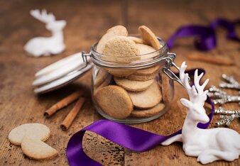 Foto von den Chai Kekserl in einem Glas. Um das Glas liegen Zimtstangen und ein violettes Band, sowie ein kleiner Porzellan Hirsch. | © SONNENTOR