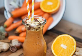 Auf dem Foto sieht man eine Karaffe mit Karotten-Orangen Smoothie. Darauf sind viele Nüsse zu sehen. Rundherum sieht man Orangen und Karotten. | © SONNENTOR