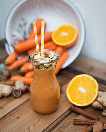 Auf dem Foto sieht man eine Karaffe mit Karotten-Orangen Smoothie. Darauf sind viele Nüsse zu sehen. Rundherum sieht man Orangen und Karotten. | © SONNENTOR