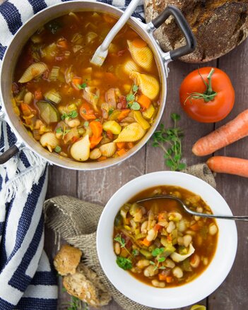 Au dem Foto sieht man einen Topf und einen Teller mit Minestrone. Daneben sieht man etwas Brot, Karotten und Tomaten. | © SONNENTOR