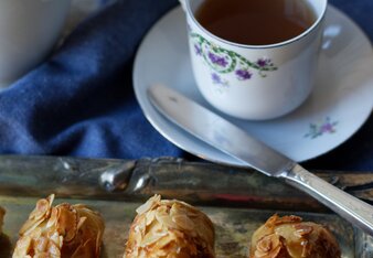 Auf dem Foto sind drei Bienenstich-Minis auf einem Silbertablett zu sehen. Dahinter steht eine Tasse Tee. | © SONNENTOR