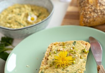 Ein Brötchen bestrichen mit dem Wildkäuterhummus und dekoriert mit einer Löwenzahnblüte liegt auf einem Teller, im Hintergrund steht die Schüssel mit dem restlichen Hummus. | © SONNENTOR