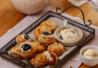 Die Scones liegen auf einem Tablett mit schwarzem Rand und schwarzen Hänkeln, ein Schälchen Clotted Cream steht ebenso darauf. Manche Scones sind mit Clotted Cream und Fruchtaufstrich dekoriert. Neben dem Tablett steht eine Teekanne und eine Tasse Tee. | © SONNENTOR