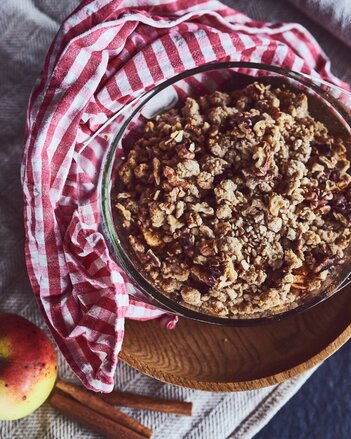 Auf dem Foto ist eine Form mit Apfel-Cranberry-Crumble zu sehen. Daneben liegen Zimtstangen und ein Apfel. | © SONNENTOR