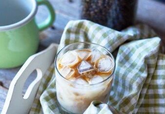 Auf dem Foto ist ein Glas mit Chai Eiskaffee zu sehen. Das Glas steht auf einem grün kariertem Küchentuch. Dahinter sind Kaffeebohnen zu sehen. | © SONNENTOR