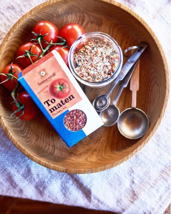 Auf dem Foto ist eine große Holzschale zu sehen, in der eine Packung Tomatenflocken, Tomaten und ein Schälchen mit mediterraner Gewürzmischung liegen. | © SONNENTOR