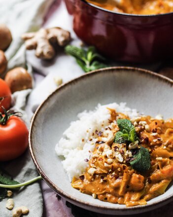 Auf dem Foto ist ein Teller mit Cashew-Curry zu sehen. Daneben ist auch Reis zu sehen. Im Hintergrund sieht man Ingwer und Tomaten. | © SONNENTOR