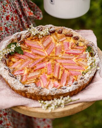 Auf dem Foto sieht man eine Rhabarber Tarte auf einem Holzbrett. Die Tarte ist mit frischen Blüten garniert. | © SONNENTOR