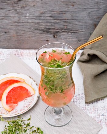 Ein bauchiges Cocktailglas ist gefüllt mit orange rosa Saft mit SONNENTOR Tschin Tschin Sirup, Grapefruit und Thymian, ergänzt mit einem rosegoldenen Trinkhalm. Ein Teller mit Grapefruit-Spalten steht daneben. | © SONNENTOR