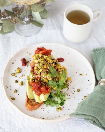 Das Frühstücksbrot ist mit Bohneneintopf, Eierspeis und Avocado belegt und mit Pistazien verziert und liegt auf einem Teller. Daneben steht eine Tasse Tee.  | © SONNENTOR