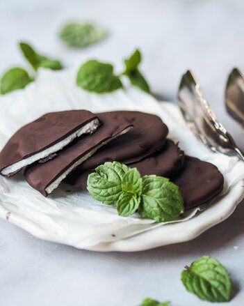 Vier Pfefferminzblättchen sind mit dunkler Schokolade überzogene Taler und liegen auf einem weißen Teller, eines ist auseinandergebrochen und man sieht die weiße Füllung. Ein Pfefferminzblatt liegt zur Garnitur daneben.  | © SONNENTOR