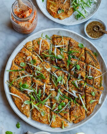 Der Kimchi Pancake ist als tellergroßer Pancake gebacken worden und in kleine Rauten geschnitten. Dekoriert ist er mit länglich geschnittenen Jungzwiebel Streifen und Sesam.  | © SONNENTOR