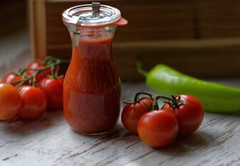 Auf dem Foto sieht man ein Glas voll mit Ketchup. Daneben sind frische Tomaten und einen grünen Paprika zu sehen. | © SONNENTOR