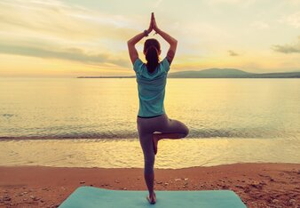 Auf dem Foto sieht man wie eine Frau am Strand die Yogaposition Der Baum eingenommen hat. | © SONNENTOR