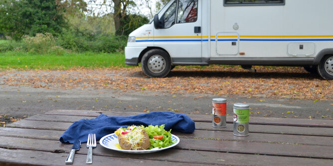 Auf dem Foto sieht man einen Tisch mit einem Teller mit Ofenkartoffel darauf. Hinter dem Tisch sieht man einen Camper. | © SONNENTOR