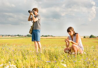 Auf dem Foto sieht man zwei Personen auf einer Wiese. Der Mann fotografiert und die Frau betrachtet Blüten. | © SONNENTOR